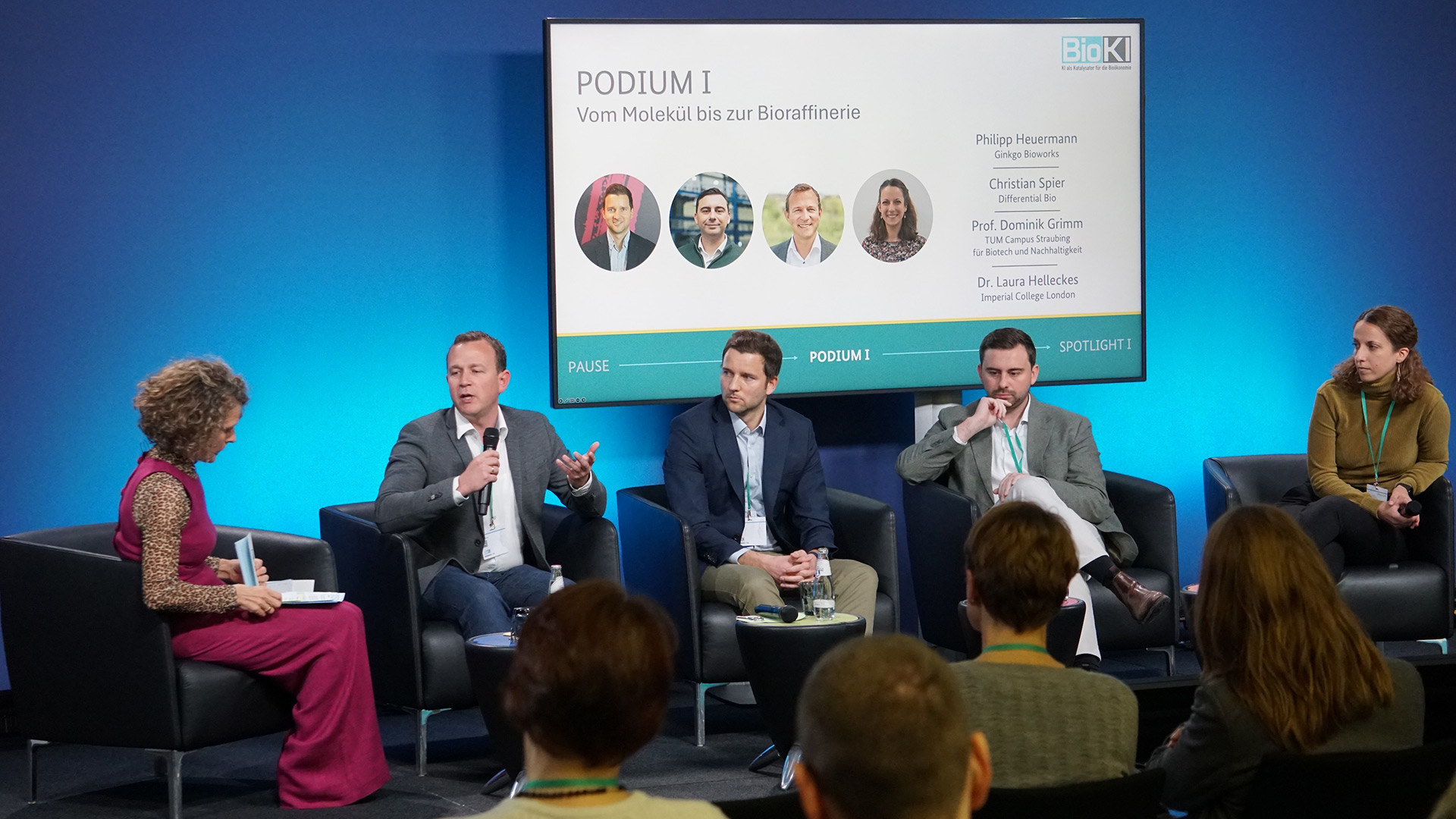 Five people, two women and three men, sitting in front of a blue background at a podium discussion. One of the men, Prof. Grimm, is holding a microphone in is hand and is speaking.